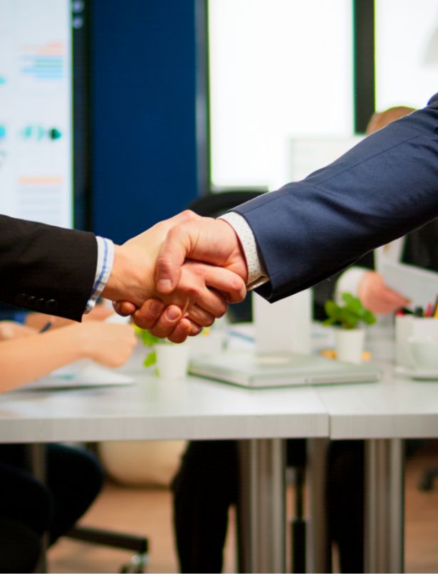 Two people in business attire shaking hands in an office setting, with a desk, documents, and blurred colleagues visible in the background.