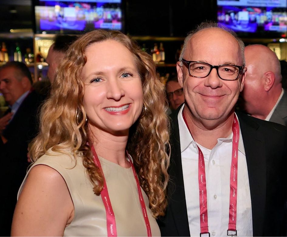 A woman with curly blonde hair and a man wearing glasses are smiling at the camera at a social event. Both are wearing name badges on lanyards. People and bar shelves are visible in the background.