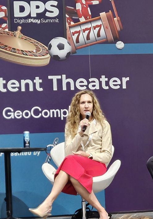 A woman with long curly hair, wearing a beige top and red skirt, sits on a white chair and speaks into a microphone at the Digital Play Summit, with gaming-themed graphics in the background.